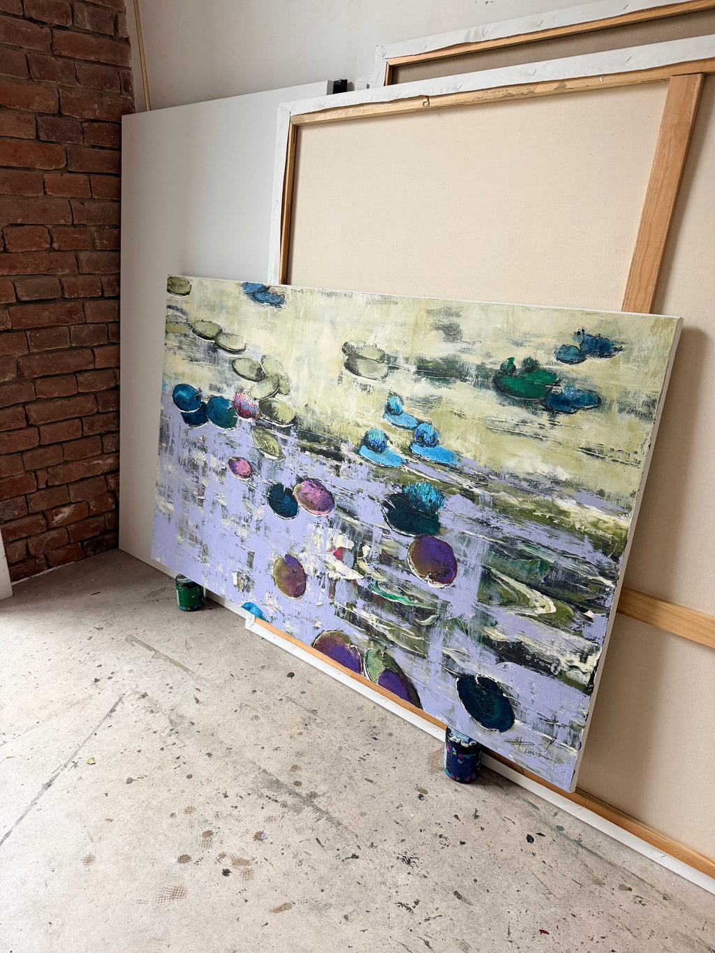 Side view of a large-format original textured water lilies painting in an artist's loft studio. The single-edition canvas artwork by Ukrainian artists is placed on the floor next to a rustic red brick wall, highlighting its authentic hand-painted texture.
